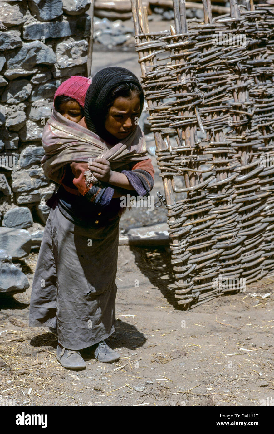 Ladakh village child girl standing carries baby back head scarf arms