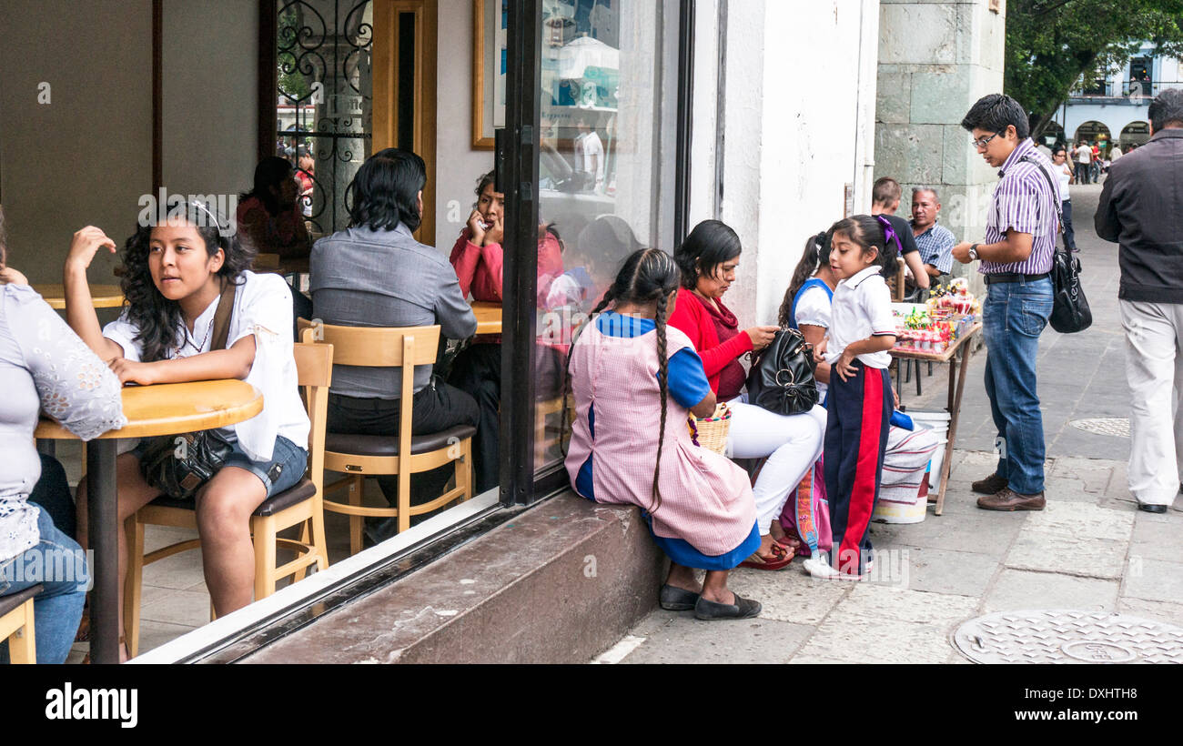 ignored by patrons inside cafe smartly dressed woman with young daughter sits on sill open window between 2 indigenous vendors Stock Photo