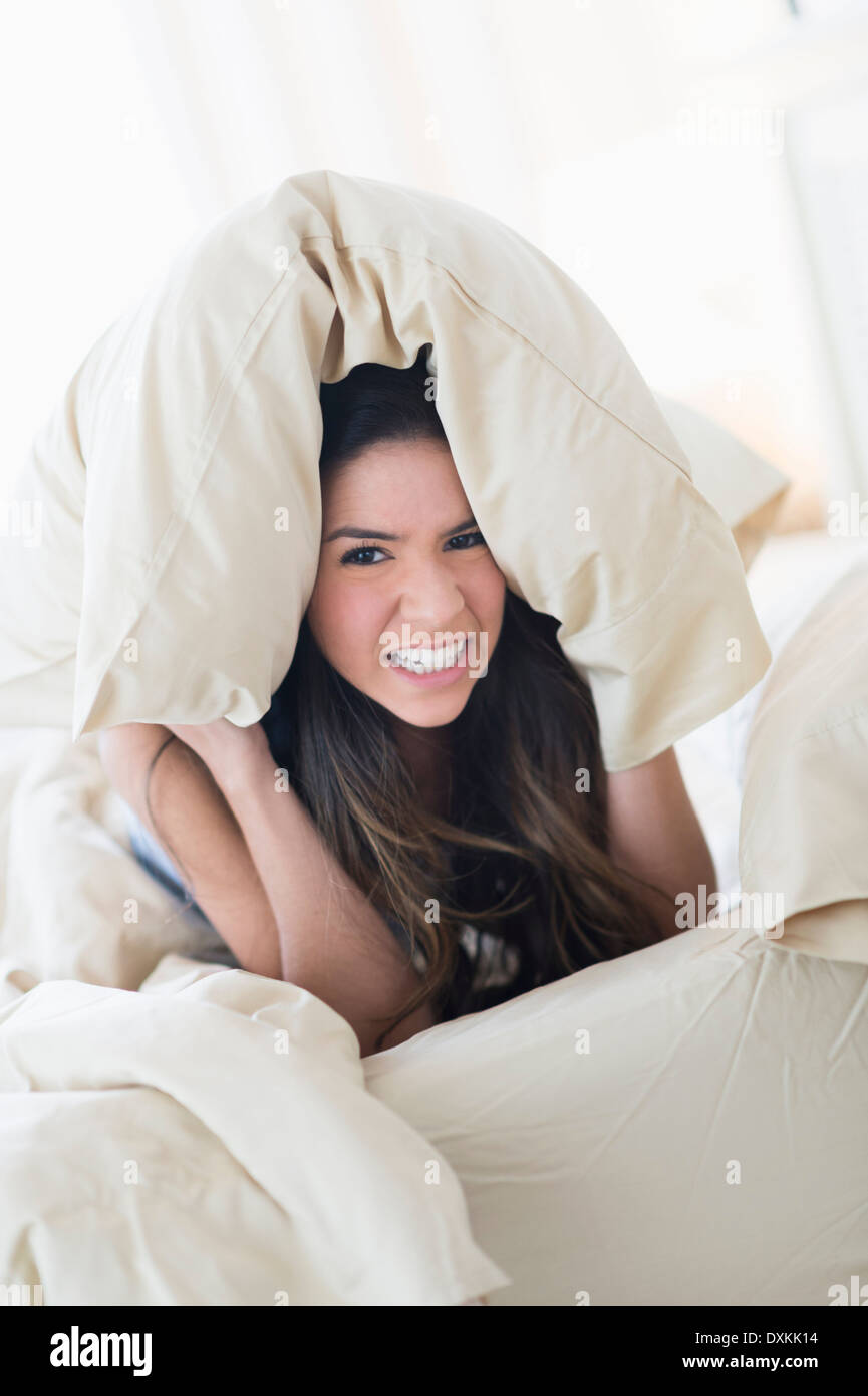 Portrait of frustrated Hispanic woman under pillow in bed Stock Photo