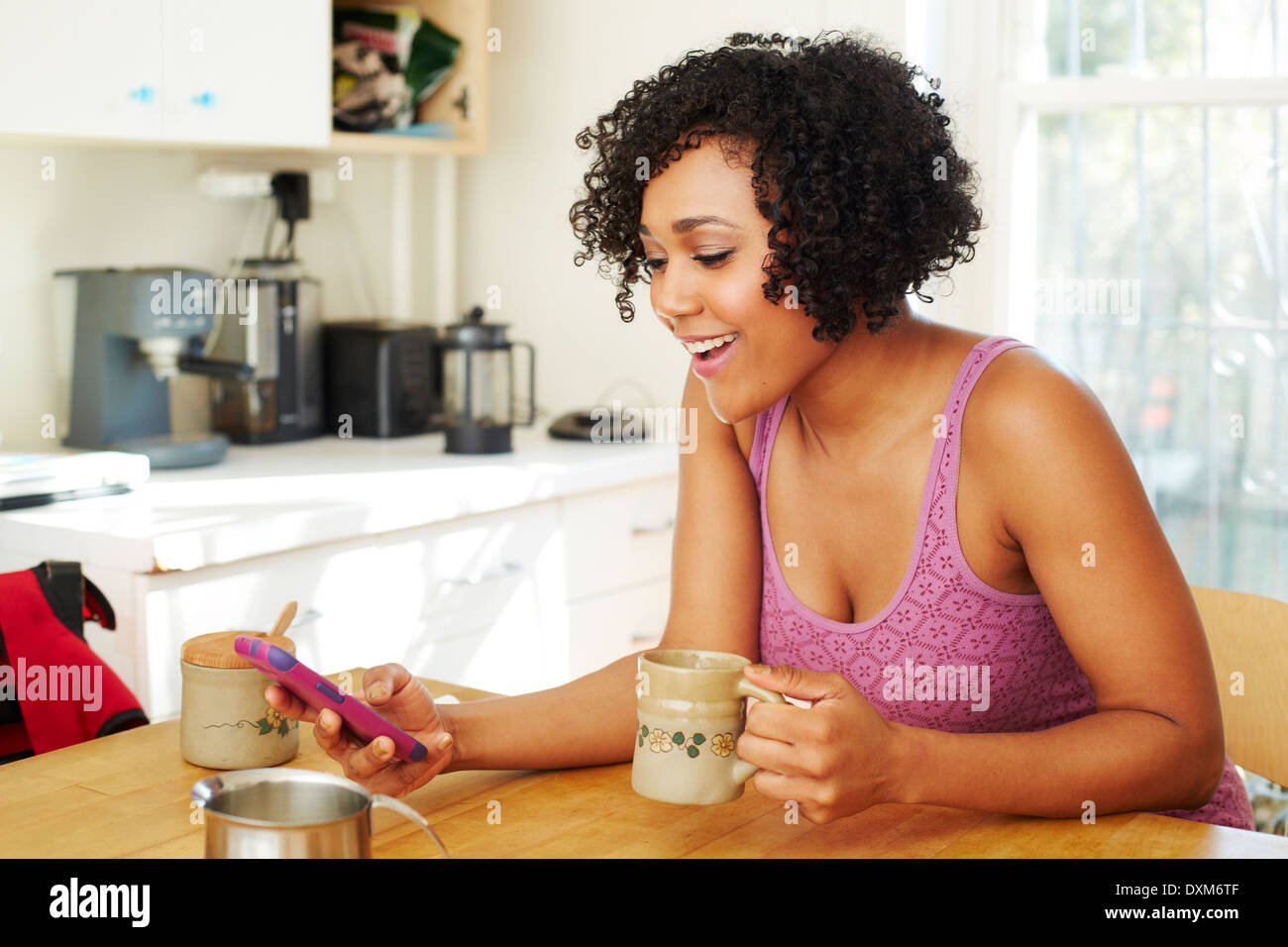 Mixed race woman using cell phone and drinking coffee in kitchen Stock Photo