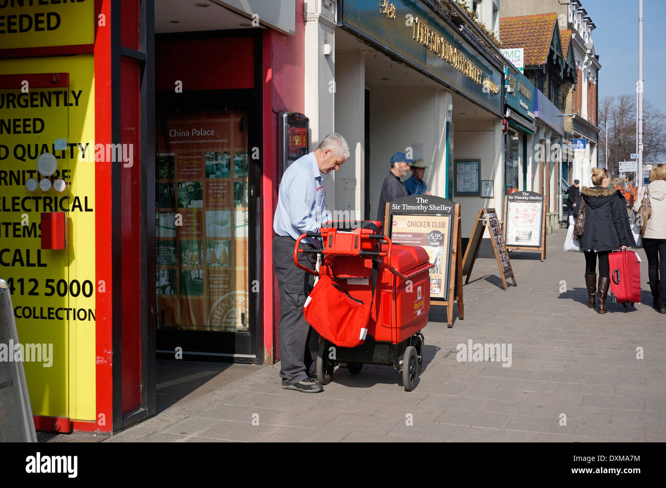 Postman on his round delivering letters & parcels using a 'high capacity' trolley (replacing the old fashioned bicycle) Stock Photo