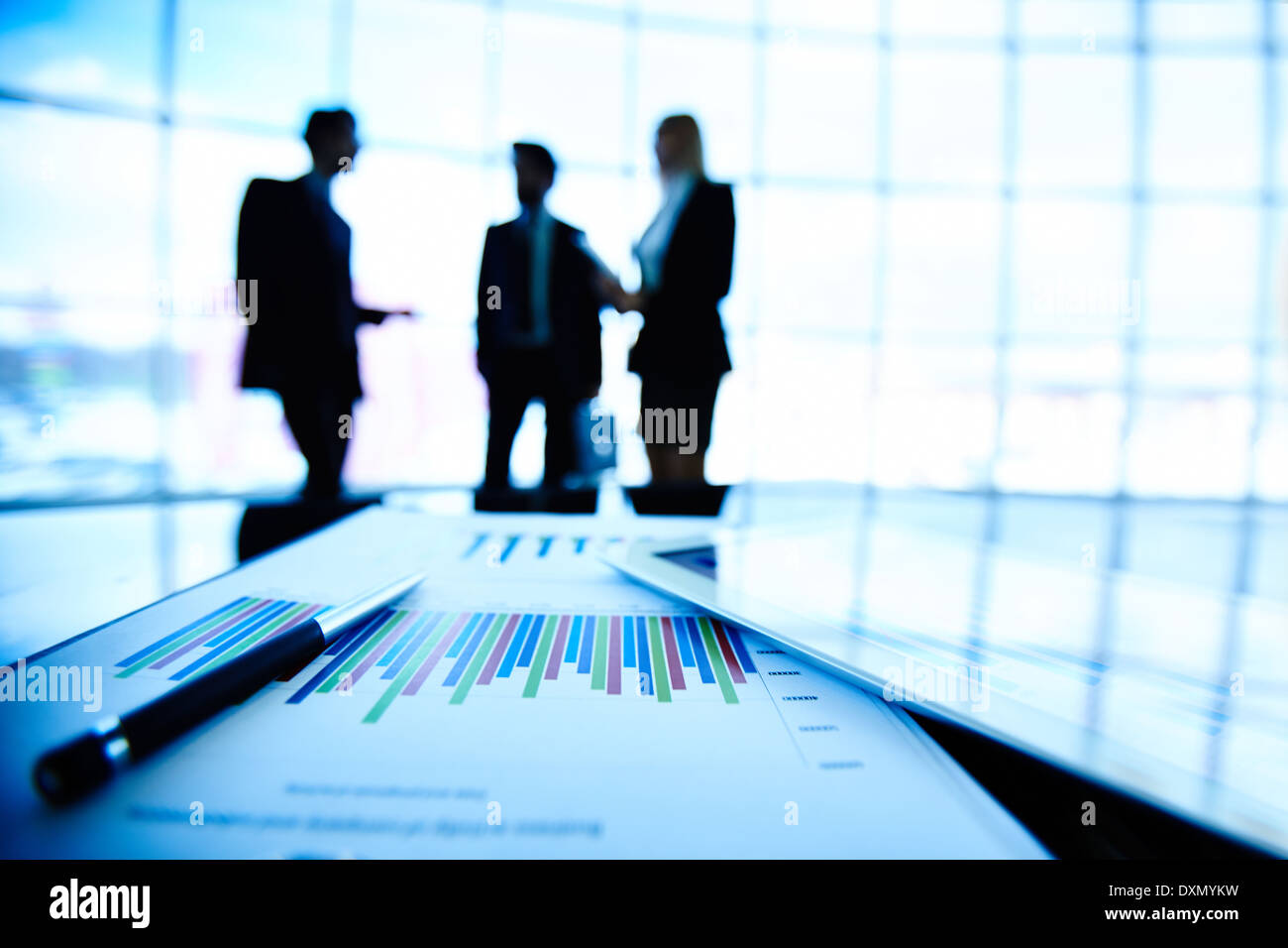 Digital tablet and financial document with pen at workplace on background of three business partners interacting Stock Photo