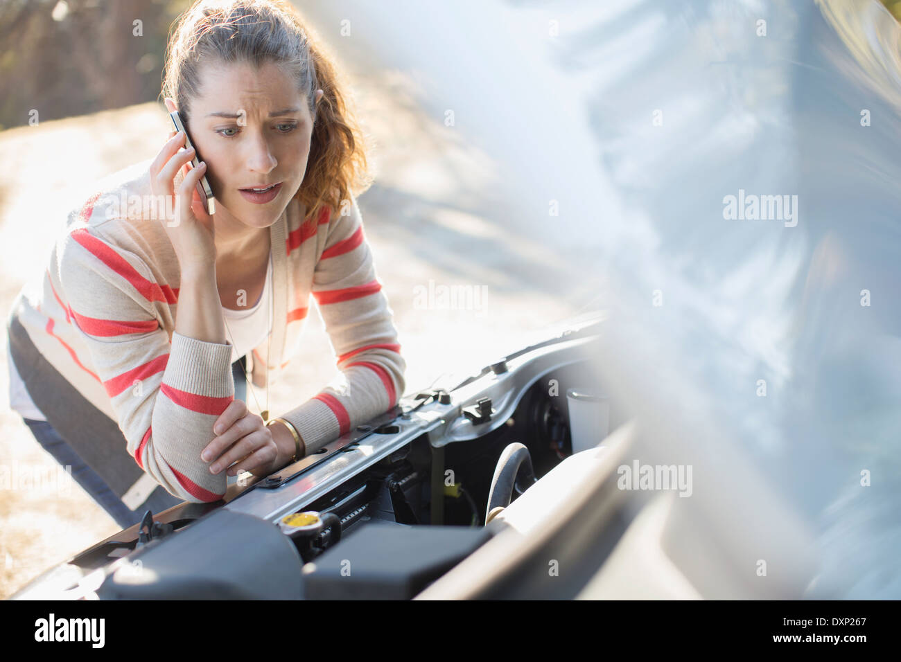 Frustrated woman talking on cell phone and looking at car engine at roadside Stock Photo