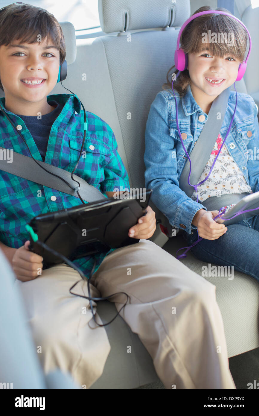 Portrait of happy brother and sister with headphones using digital tablets in back seat of car Stock Photo