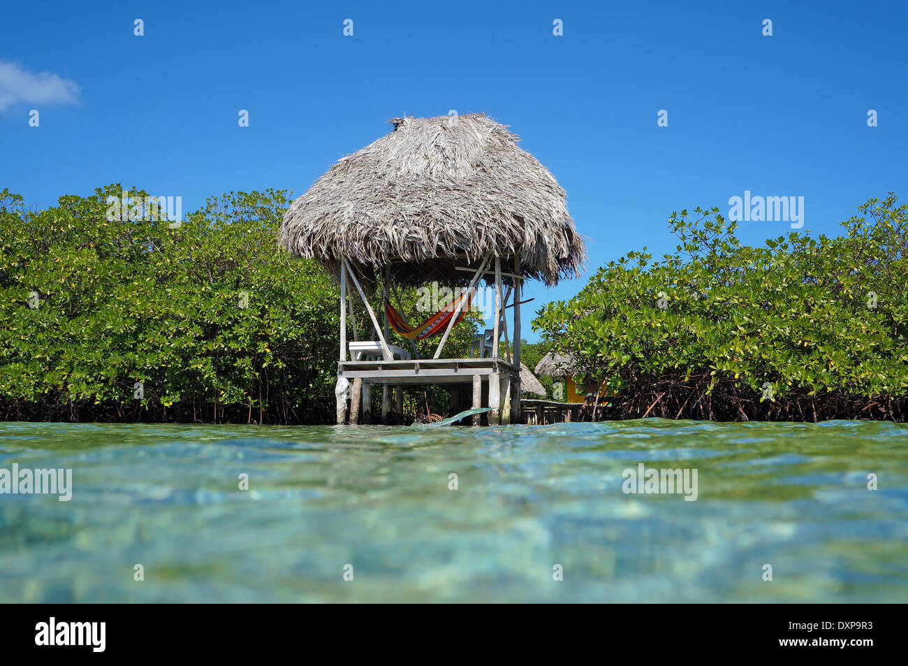 Thatched hut over water with hammock viewed from the water surface with mangrove trees and a fish jumping out of the water Stock Photo