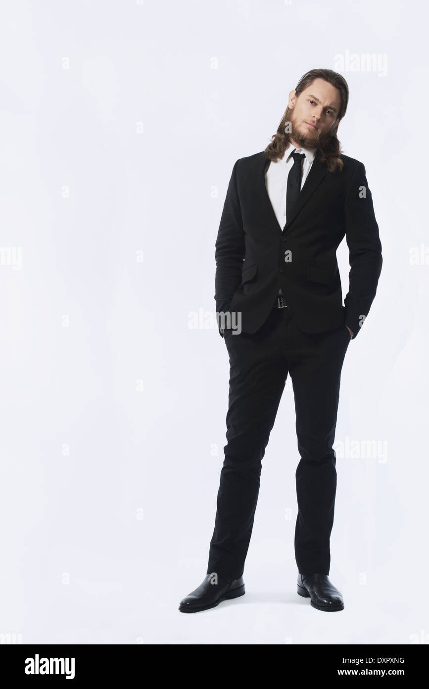 A bearded long hair man, male model standing in black suit and necktie,  hands in pocket posing Stock Photo - Alamy