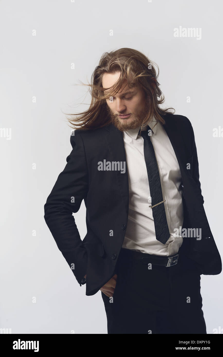 A bearded man, male model, long hair posing in a black suit and necktie looking down with hair movement, a men's fashion concept Stock Photo