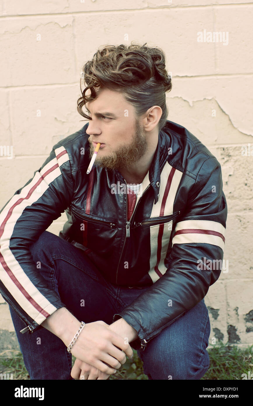 A bearded man in pompadour vintage hairstyle smoking a cigarette squatting down, an edgy vintage concept Stock Photo