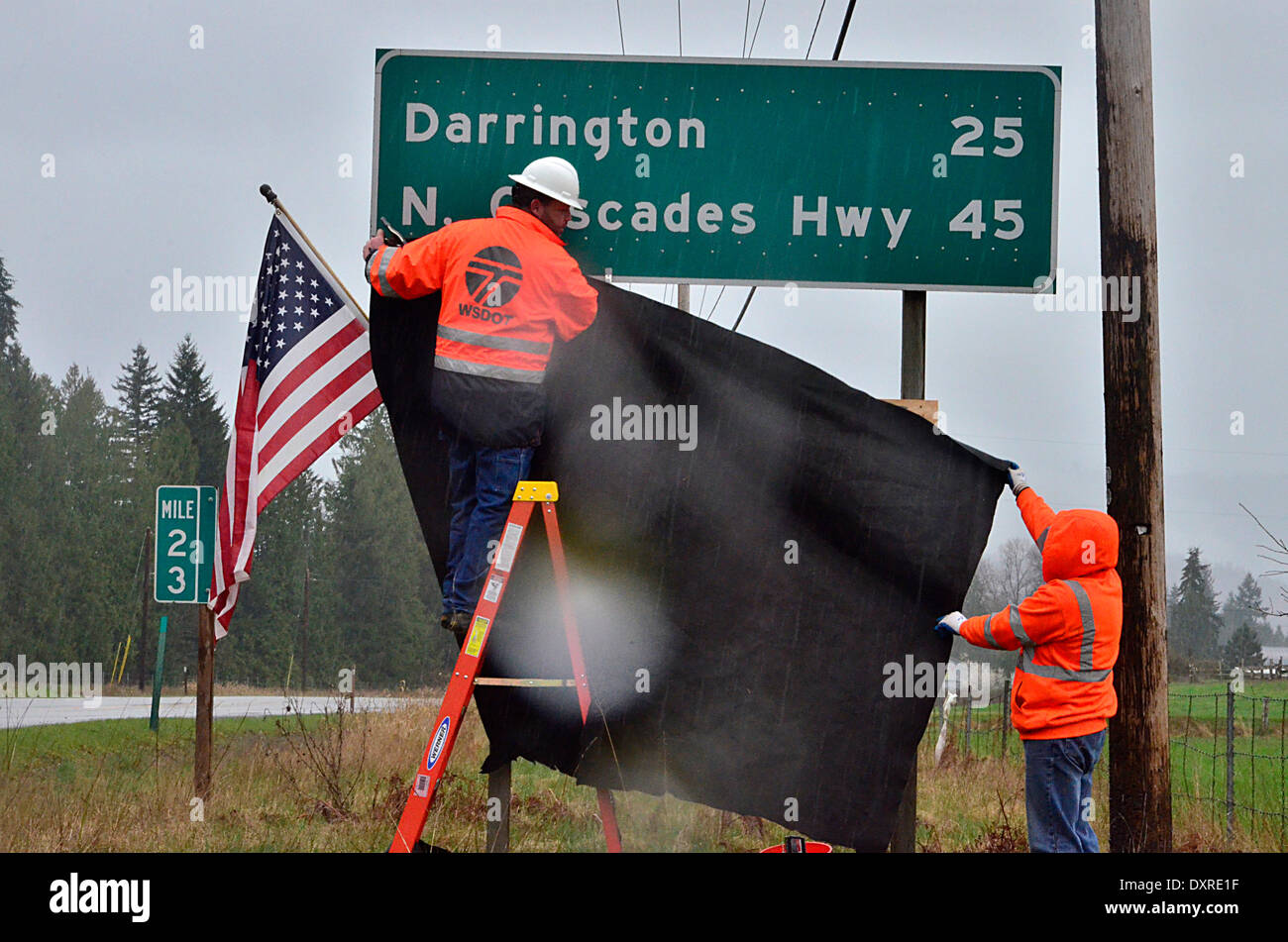 Washington State Department of Transportation workers hang an American flag and draped black material under the Darrington North Cascades Highway sign on route 530 west of the area where a massive landslide killed at least 28 people and destroyed a small riverside village in northwestern Washington state March 29, 2014 in Oso, Washington. Stock Photo