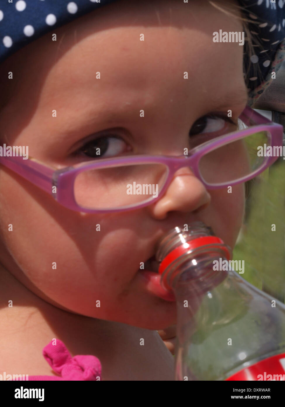 Child wearing pink glasses and a sun hat takes a drink of 'pop' at a local village fete whilst waiting for an event to start. Stock Photo