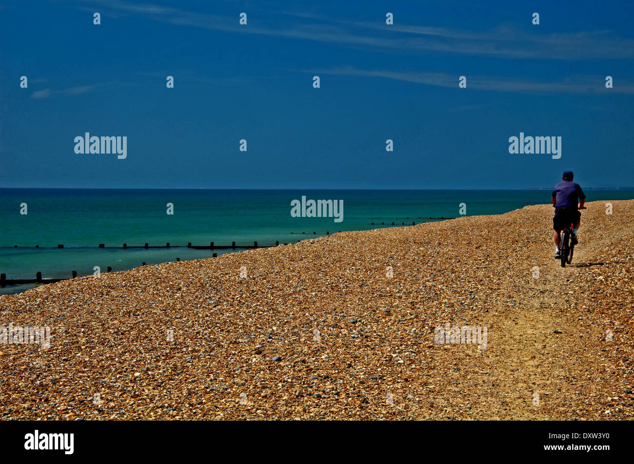 A cyclist travels away from the camera riding along the shingle beach at Climping Gap near Littlehampton. Stock Photo