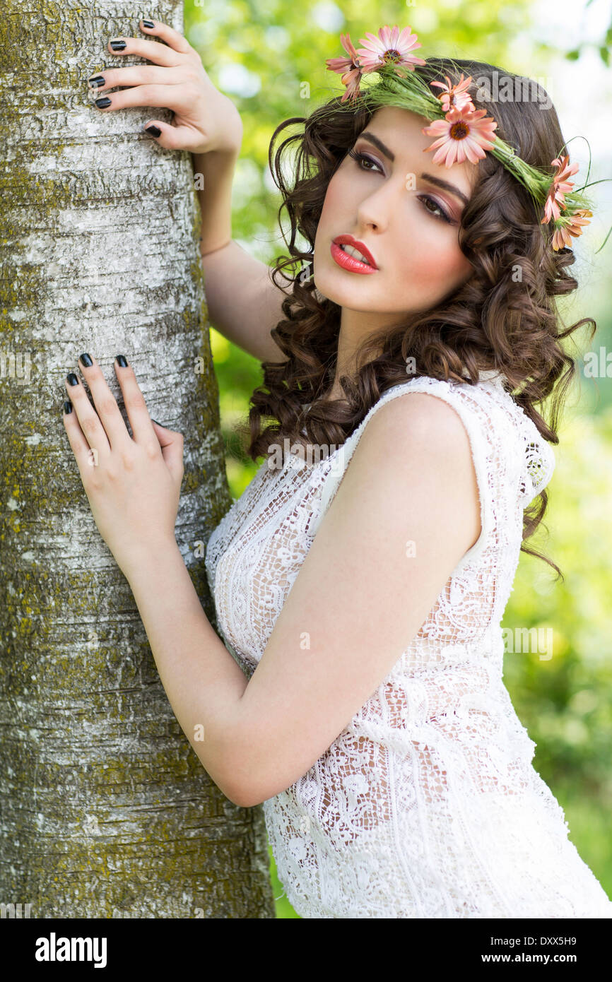 Young woman with flower wreath as headdress poses at a tree trunk Stock Photo