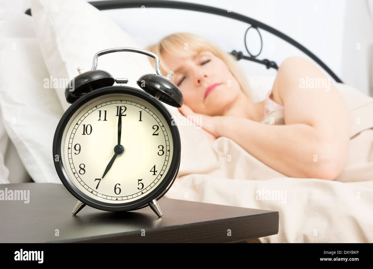 A middle age woman sleeps while alarm clock in foreground reads seven o'clock. Stock Photo