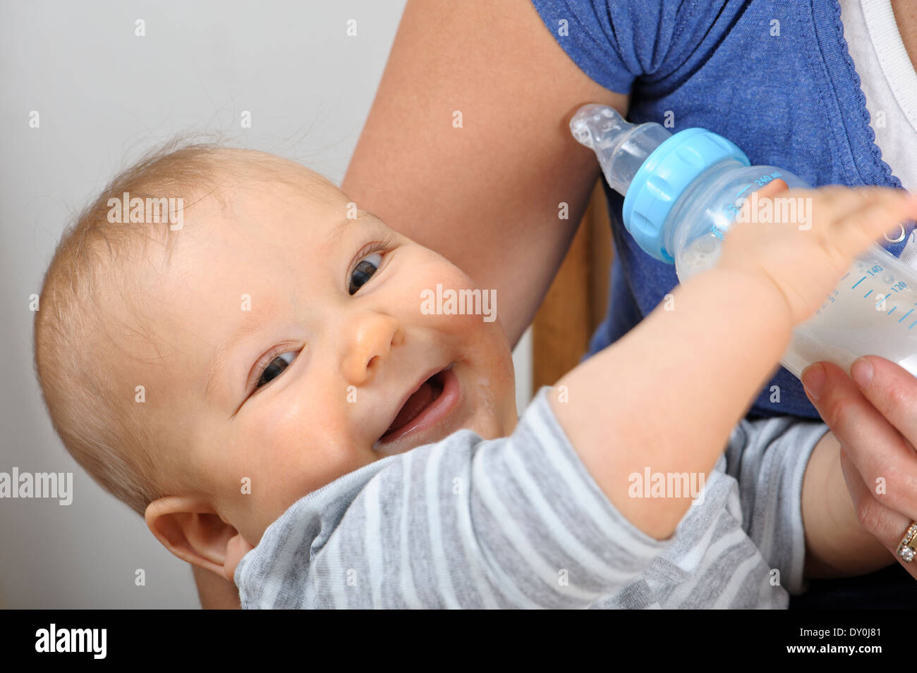 Baby bottle feeding milk in his mother's arms. He is looking at the camera and smiling Stock Photo