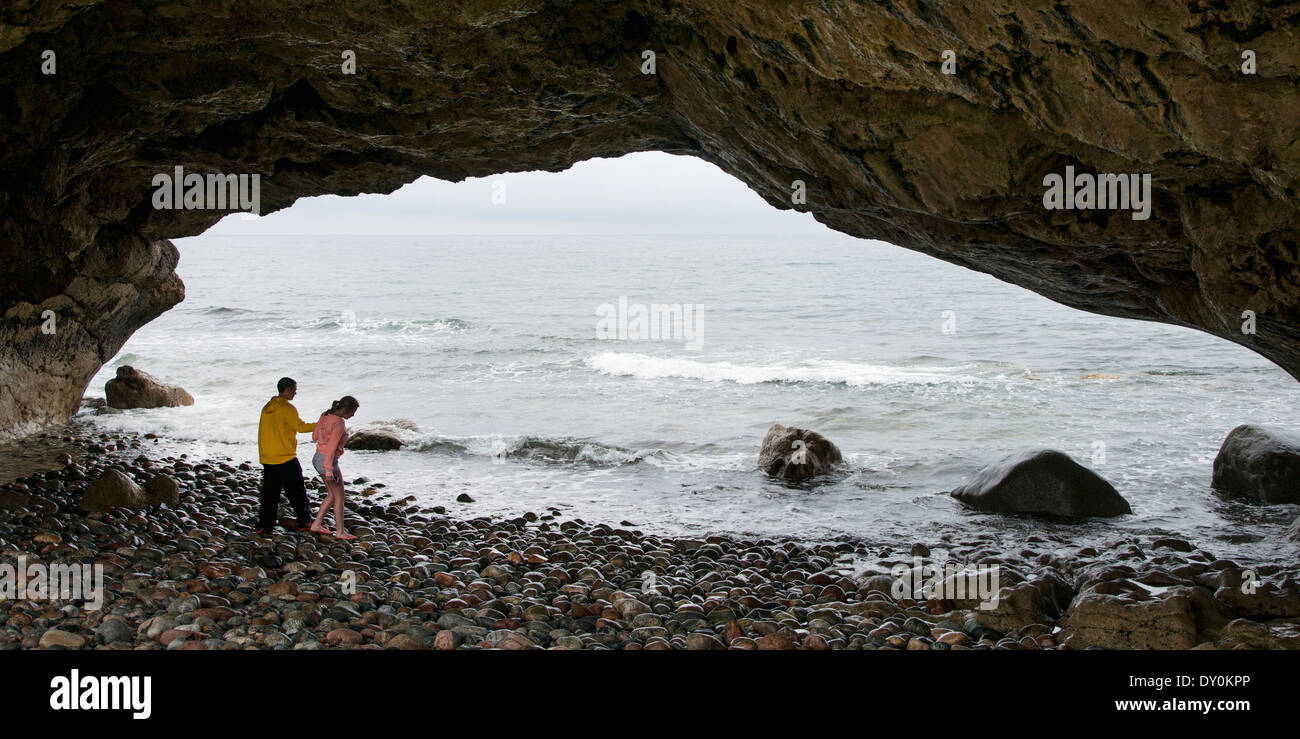Tourists walk on the rocky beach under an arched natural rock formation at the water's edge; Portland Creek, Canada Stock Photo
