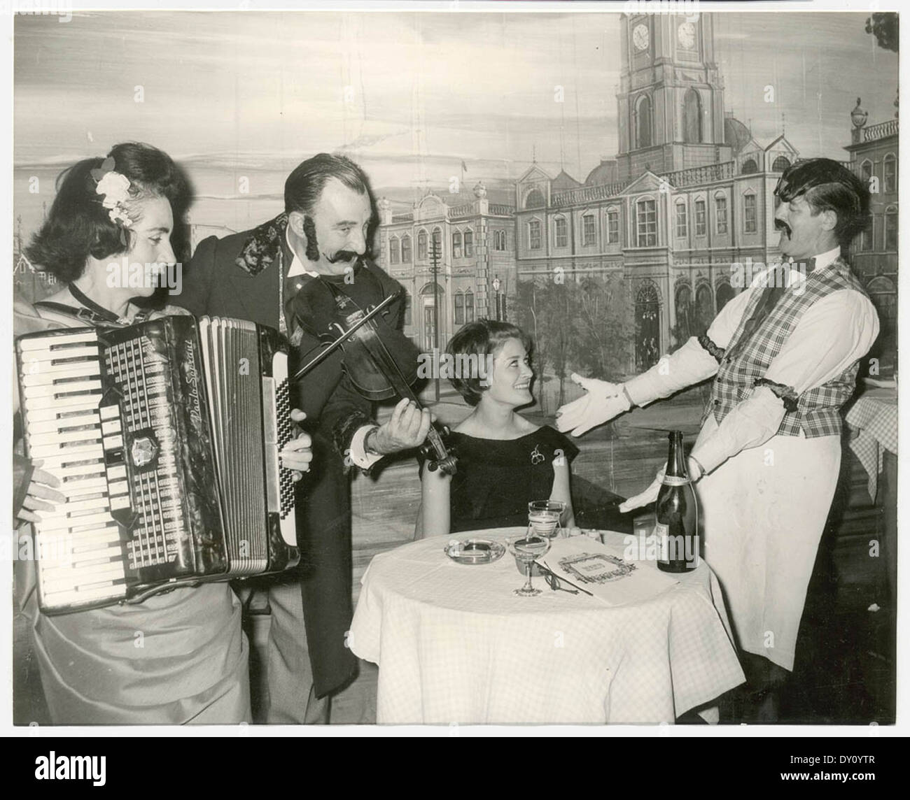 This photograph captures Elizabeth Connelly, a TWA air hostess from New York, being serenaded at the Music Hall Restaurant in April 1965. The scene features the head waiter alongside Mr. and Mrs. George Miller, who are playing the violin and accordion. Stock Photo