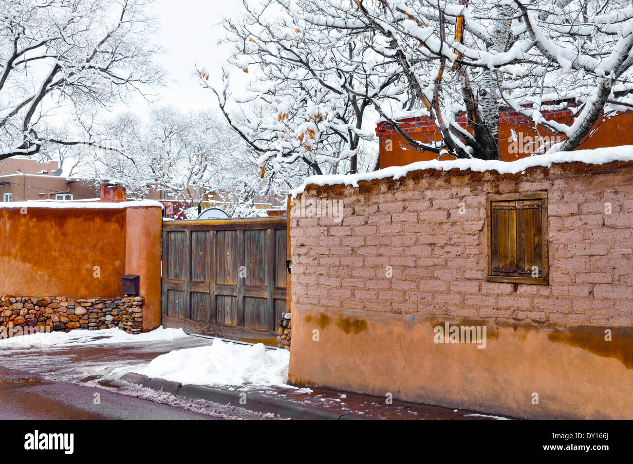 A winter's snow scene features old and new adobe walls and wooden gate in tree laden Canyon Road area of Santa Fe, NM Stock Photo