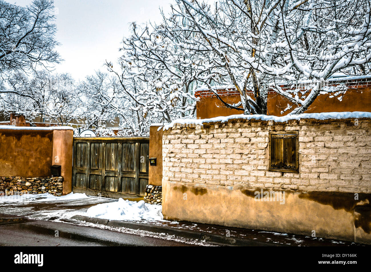 A winter snow scene features old and new adobe walls and wooden gate in tree laden Canyon Road area of Santa Fe, NM Stock Photo