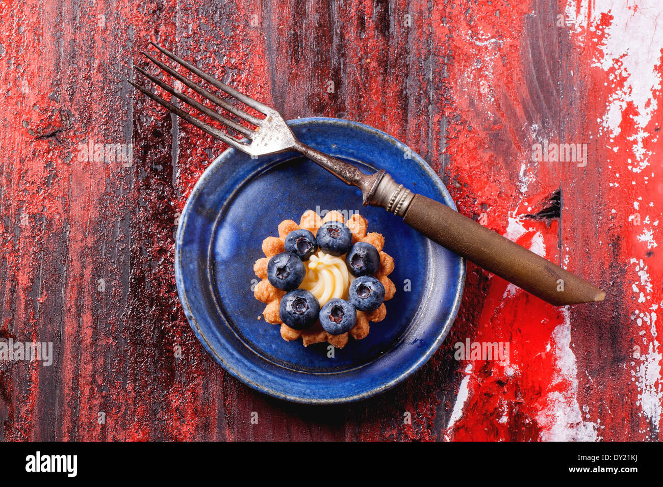 Top view on blueberry mini tart served on blue ceramic plate with vintage fork over black and red wooden background. Stock Photo