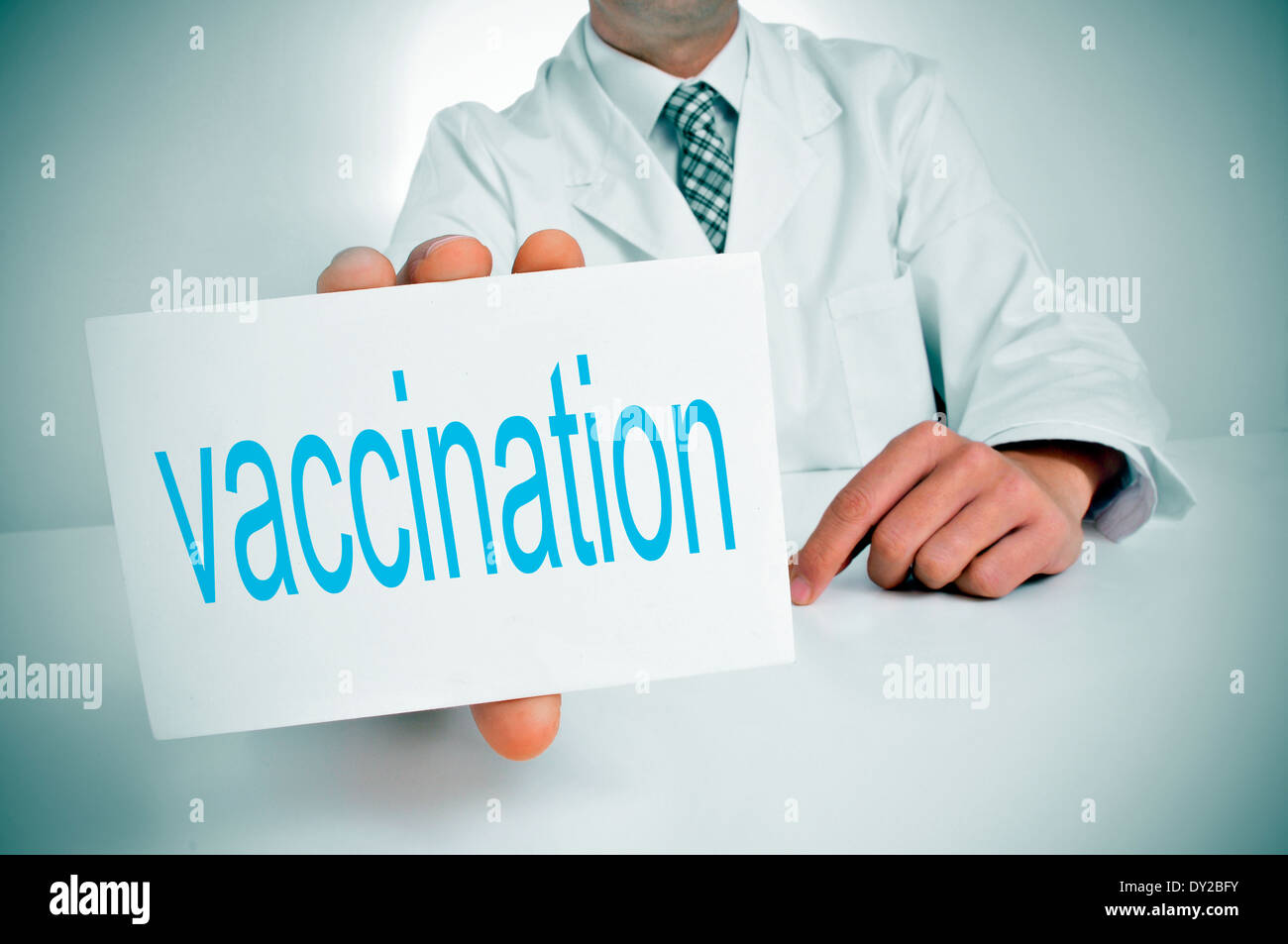 a man wearing a white coat showing sitting in a desk a signboard with the word vaccination written in it Stock Photo
