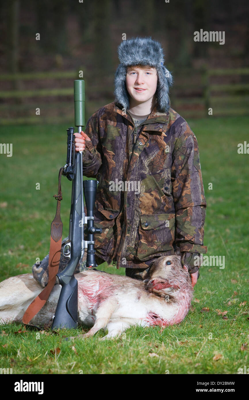 A young male deer stalker with a dead deer and a rifle in a grass field at dusk Stock Photo