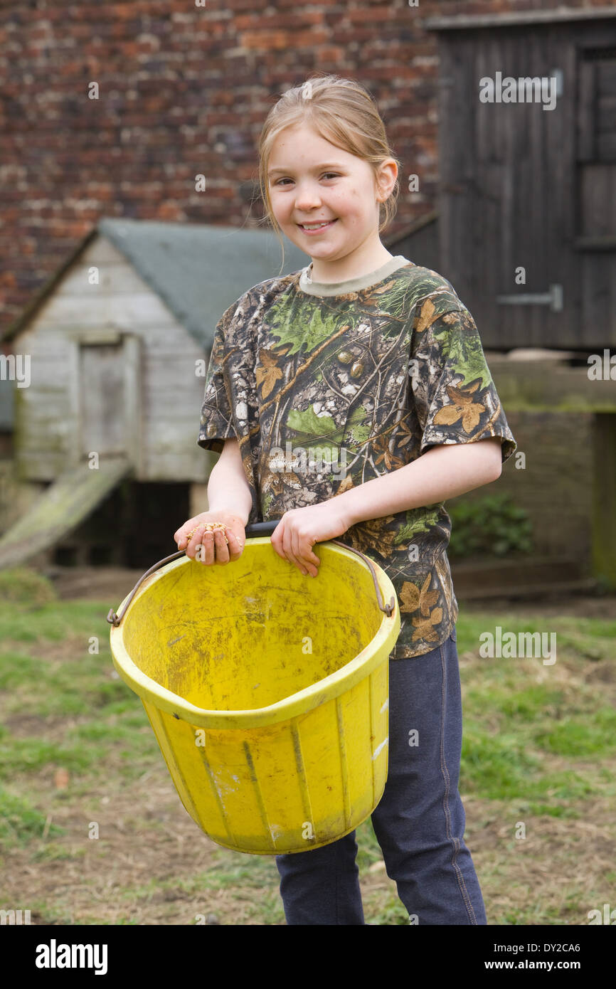 A young girl holding a bucket of chicken feed outside with a wooden hen house in the background Stock Photo