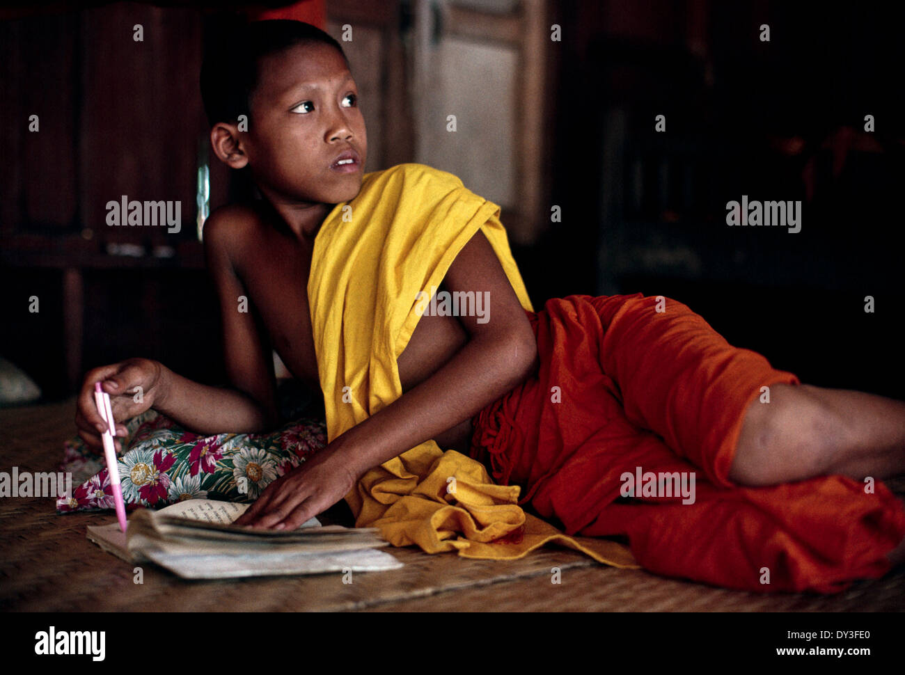 Laos young monk temple boy reclining studying homework holding pen reed mat  cotton robe dark background light foreground Stock Photo