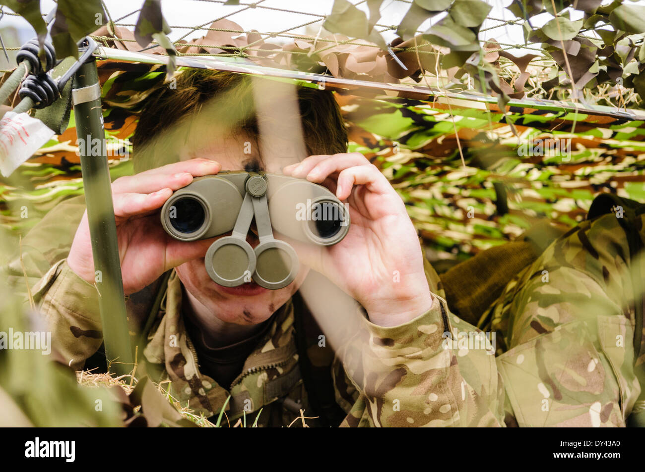 A soldier looks through binoculars as she hides underneath camouflage netting in a forward observation post Stock Photo