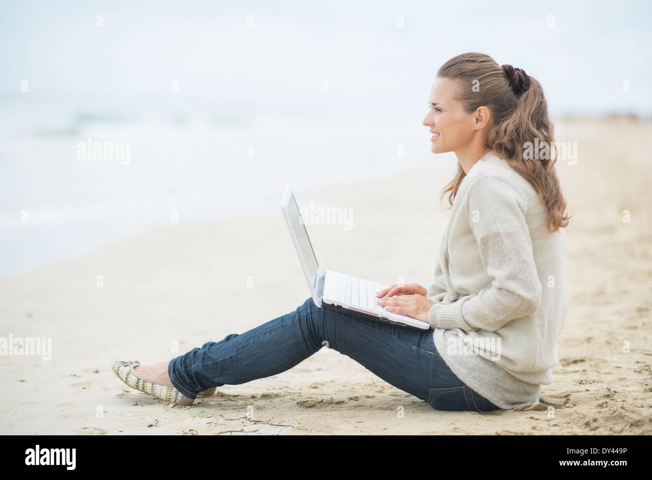 Happy young woman sitting with laptop on cold beach Stock Photo - Alamy