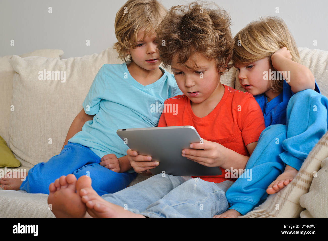 An 8-9 year old is playing a game on a touch screen tablet. His younger brothers are watching every move intently Stock Photo