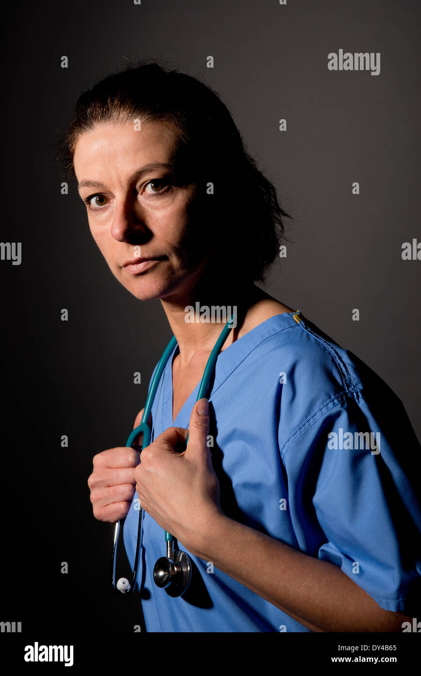 Tired looking female nurse in blue scrubs against a dark background. Stock Photo