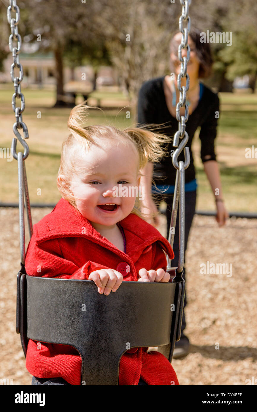 Beautiful young mother playing with adorable, cute 16 month baby girl on park playground swings Stock Photo
