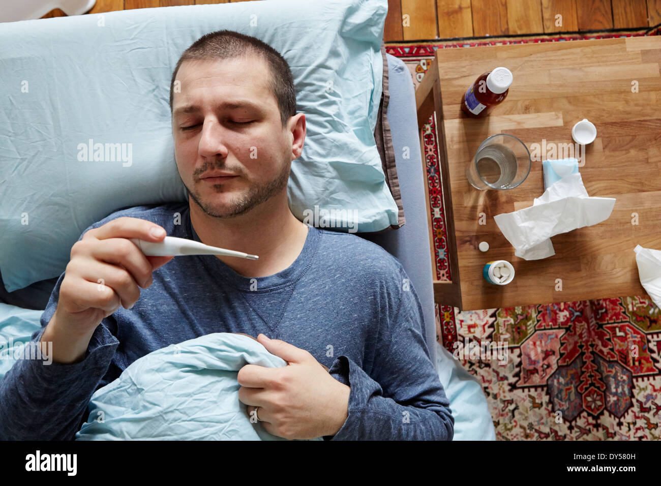 Mid adult man lying in bed looking at thermometer reading Stock Photo
