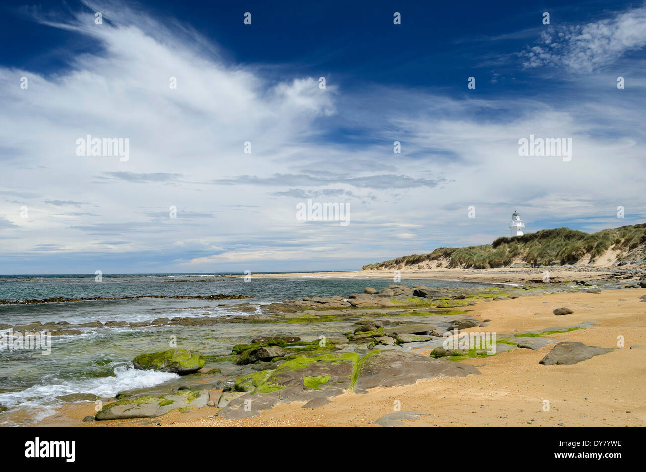 Lighthouse at Waipapa Point with clouds in the sky, sandy beach with algae covered rocks at the front, Otara, Fortrose Stock Photo