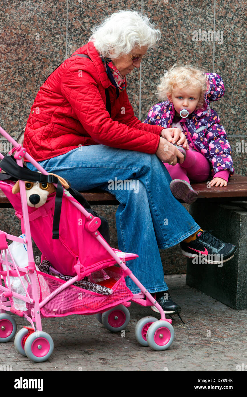 Grandmother and granddaughter senior woman and toddler young and old pink pram child pacifier girl Grandparent putting on a shoe take care of Stock Photo