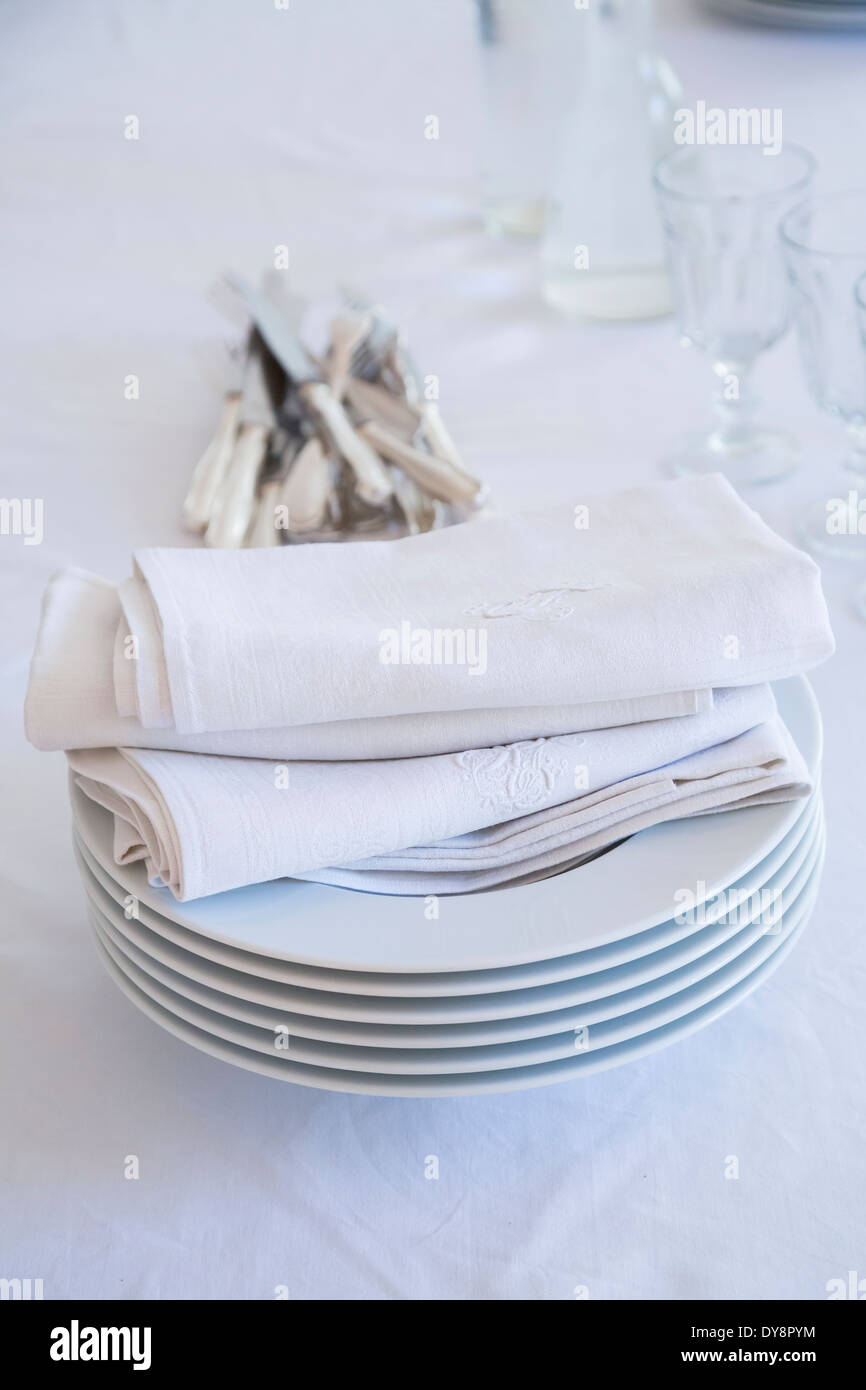 Glasses, stack of plates with cloth napkins and silver cutlery on white table cloth Stock Photo