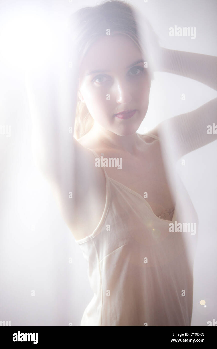 Portrait of attractive young blond woman peering through net curtains with lens flare Stock Photo