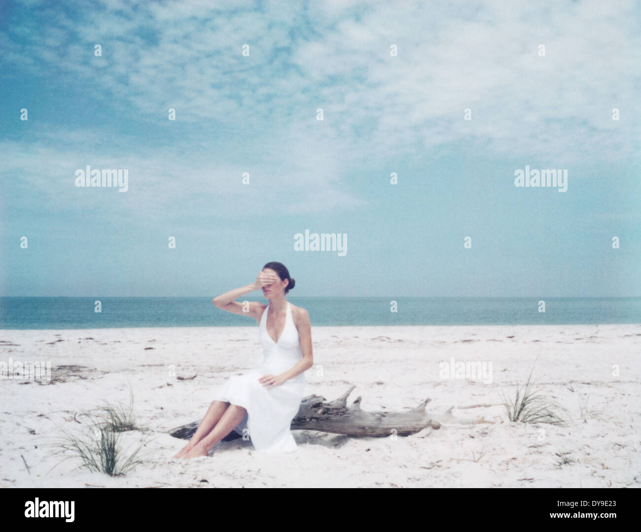 Woman sitting on driftwood at the beach, hand covering eyes Stock Photo