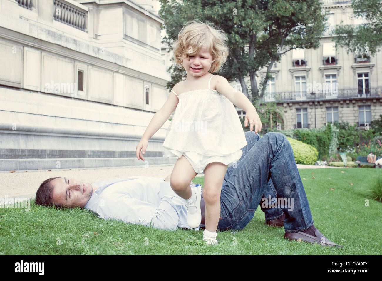 Toddler girl walking outdoors, father lying on grass in background Stock Photo