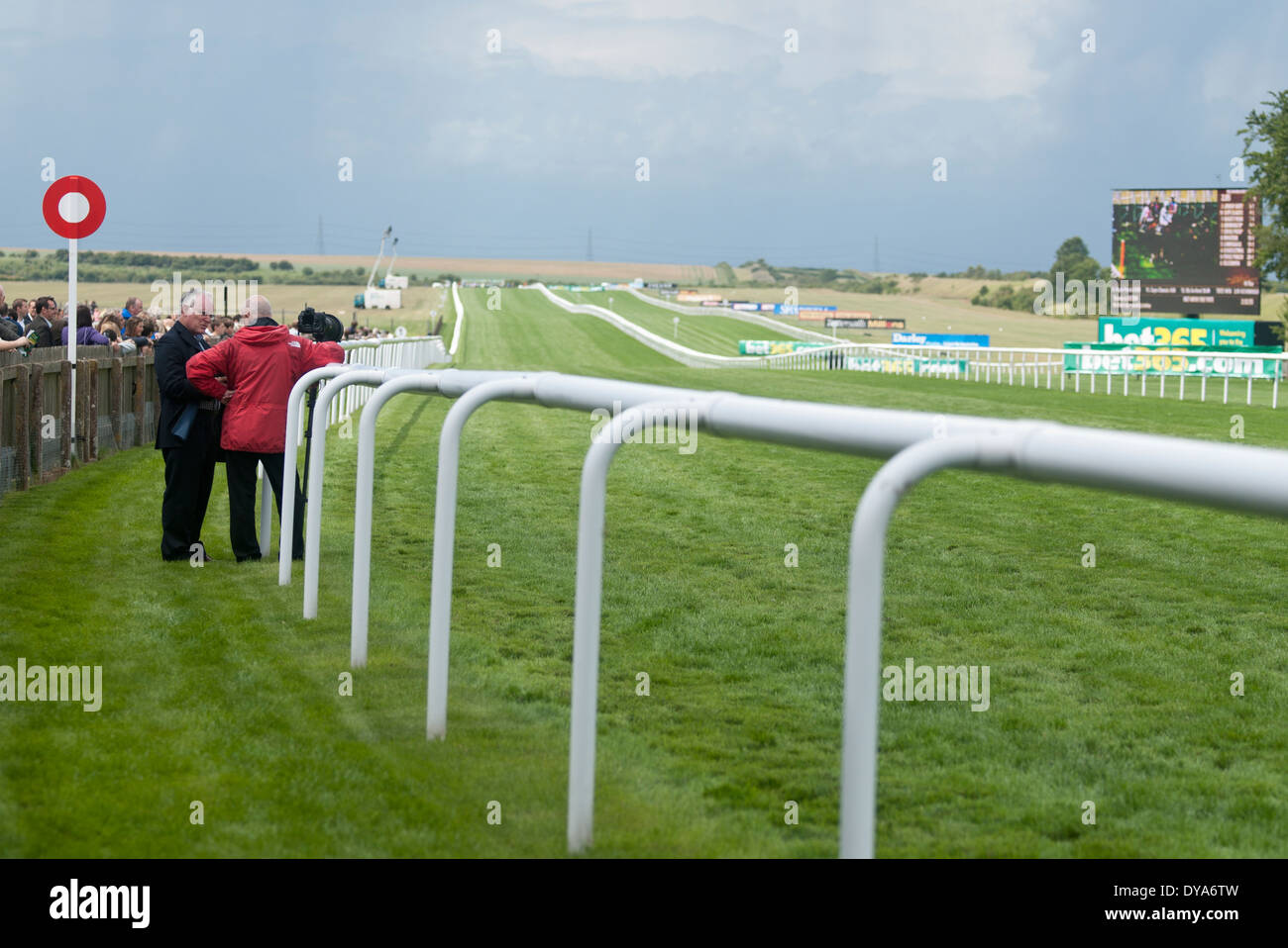 group of bystanders next to a race course waiting for horses to pass, red jacket green lawn, white fence, cloudy sky Stock Photo