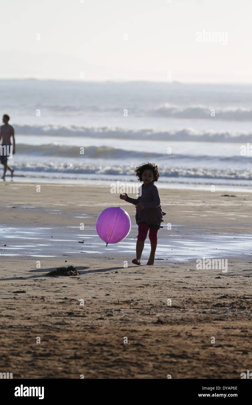 A girl runs from the waves on a beach in Essaouira, Morocco one late evening with a bright purple balloon trailing behind her Stock Photo