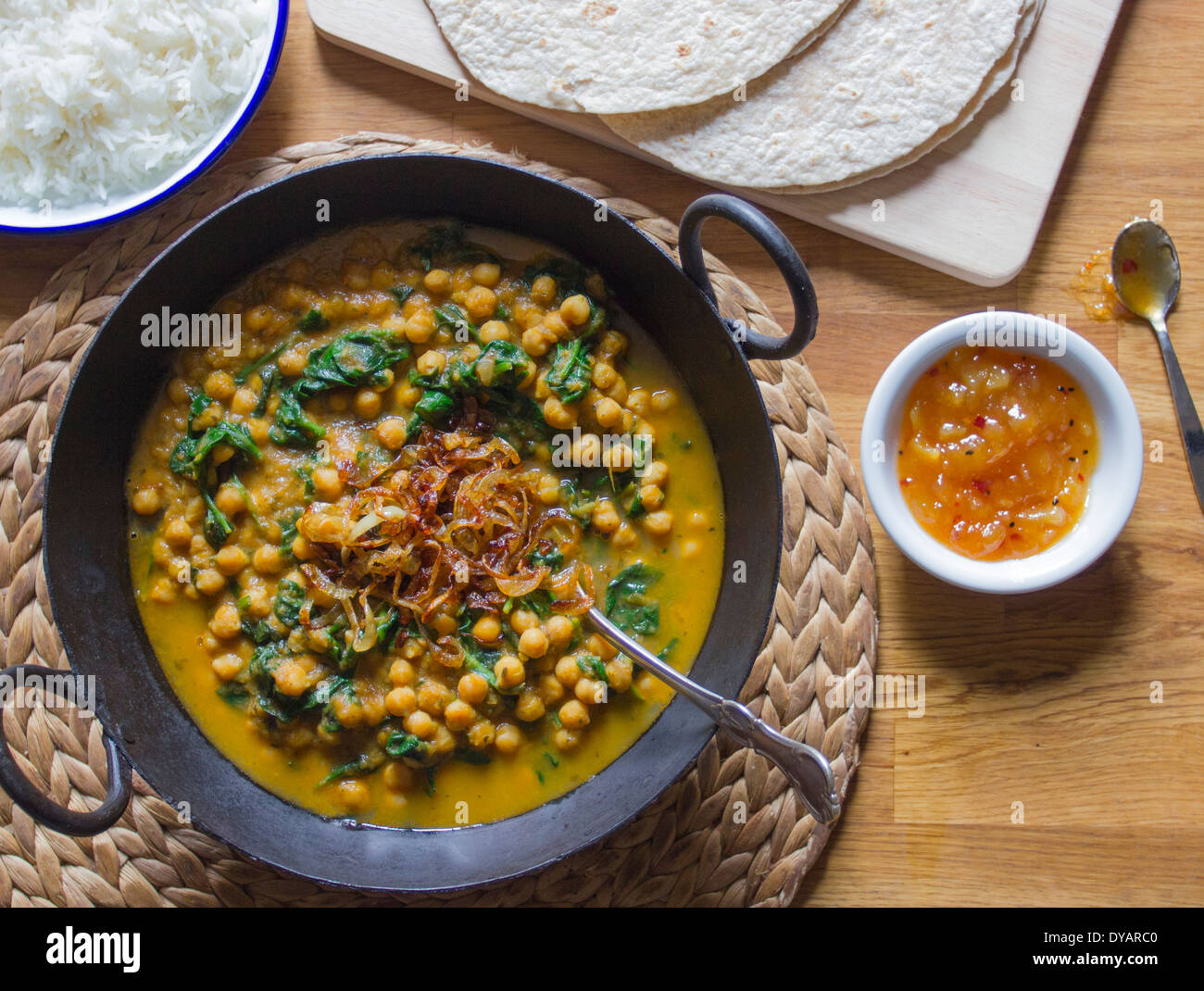 Chickpea and spinach curry with caramelised onions, rice, chapatis and mango chutney on an oak table Stock Photo