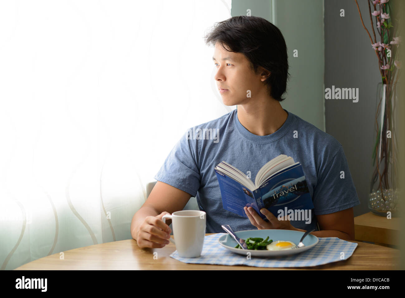 Asian man holding a book having breakfast, sitting and looking out the window daydreaming, thinking. Stock Photo