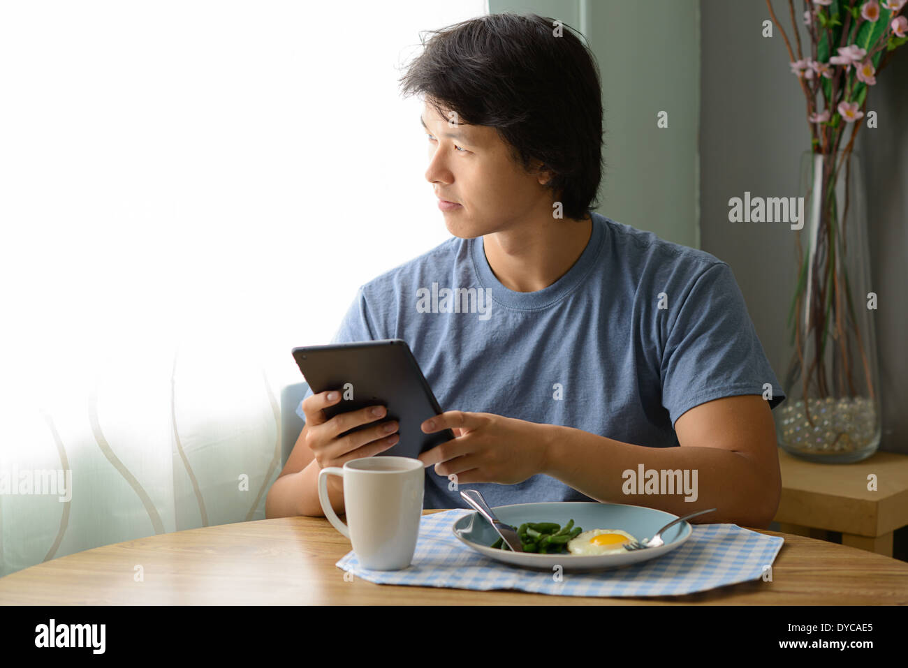Asian man holding an ipad having breakfast, sitting and looking out the window daydreaming, thinking. Stock Photo