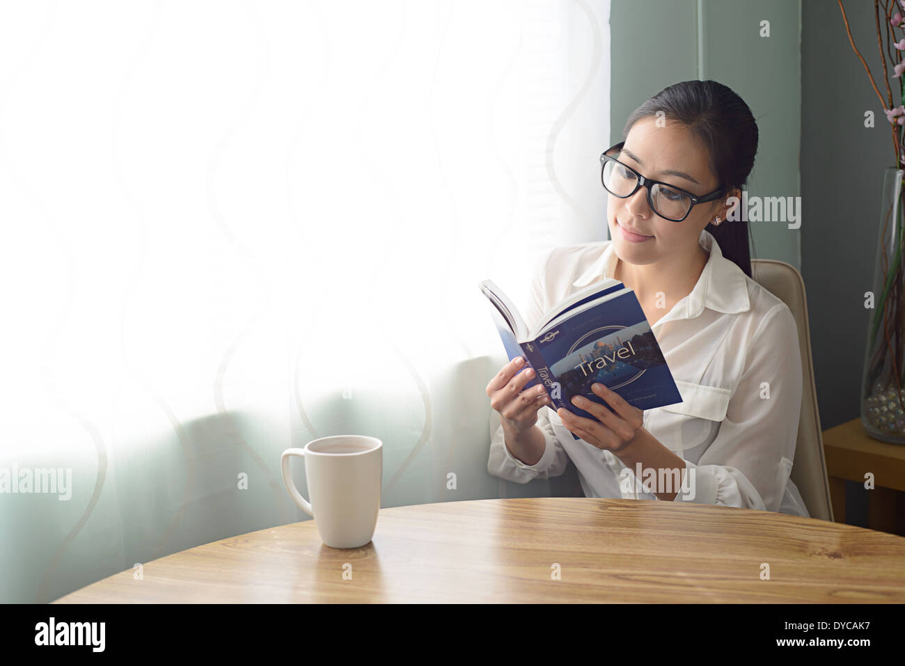 A beautiful young Asian woman in ponytail sits in her home, wearing eyeglasses reading a travel book smiling. Stock Photo