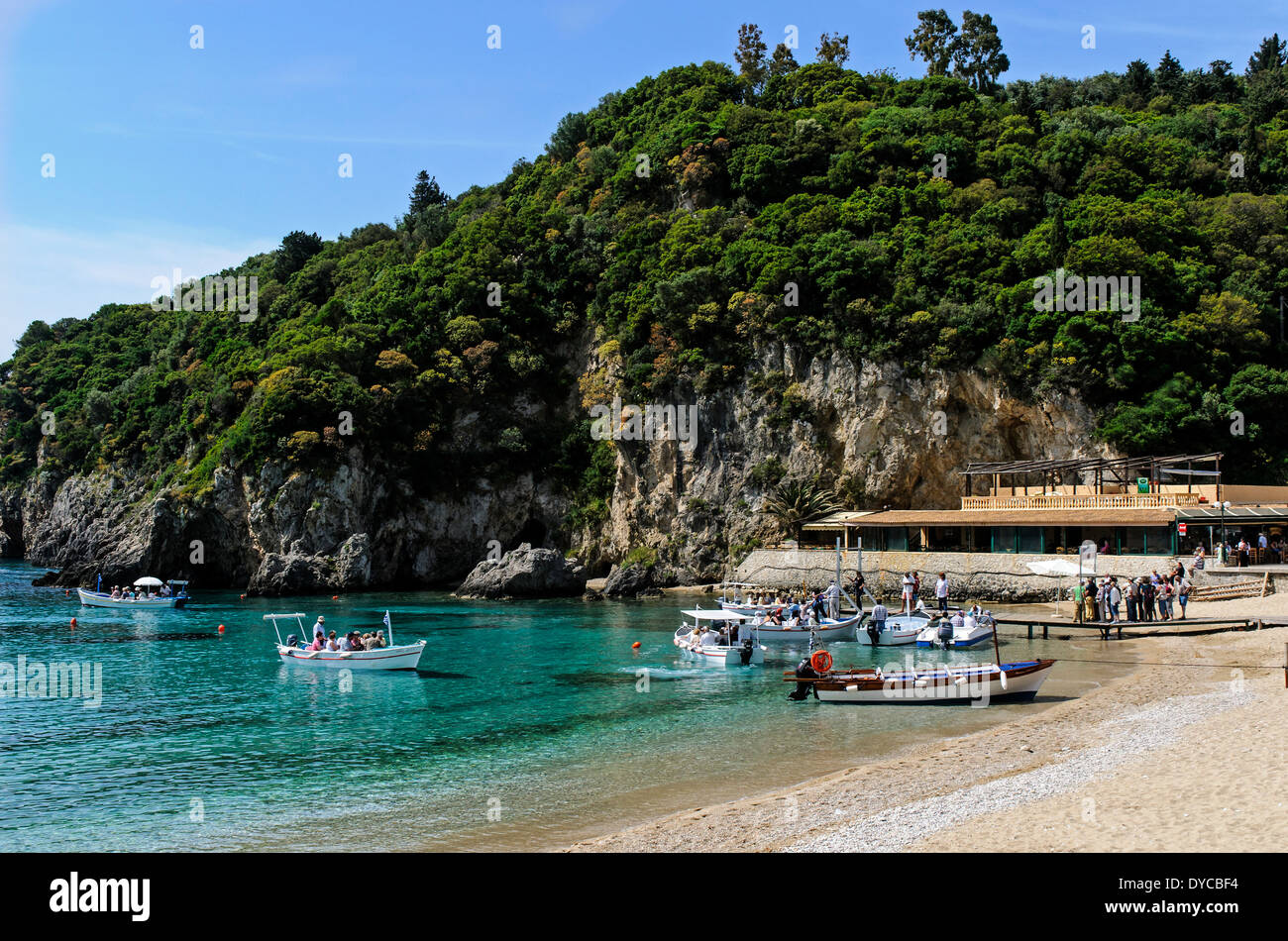 Tourists queue by the rock face of the verdant hillside for a boat trip on the   turquoise water of Agios Spyridon Bay, Corfu Stock Photo