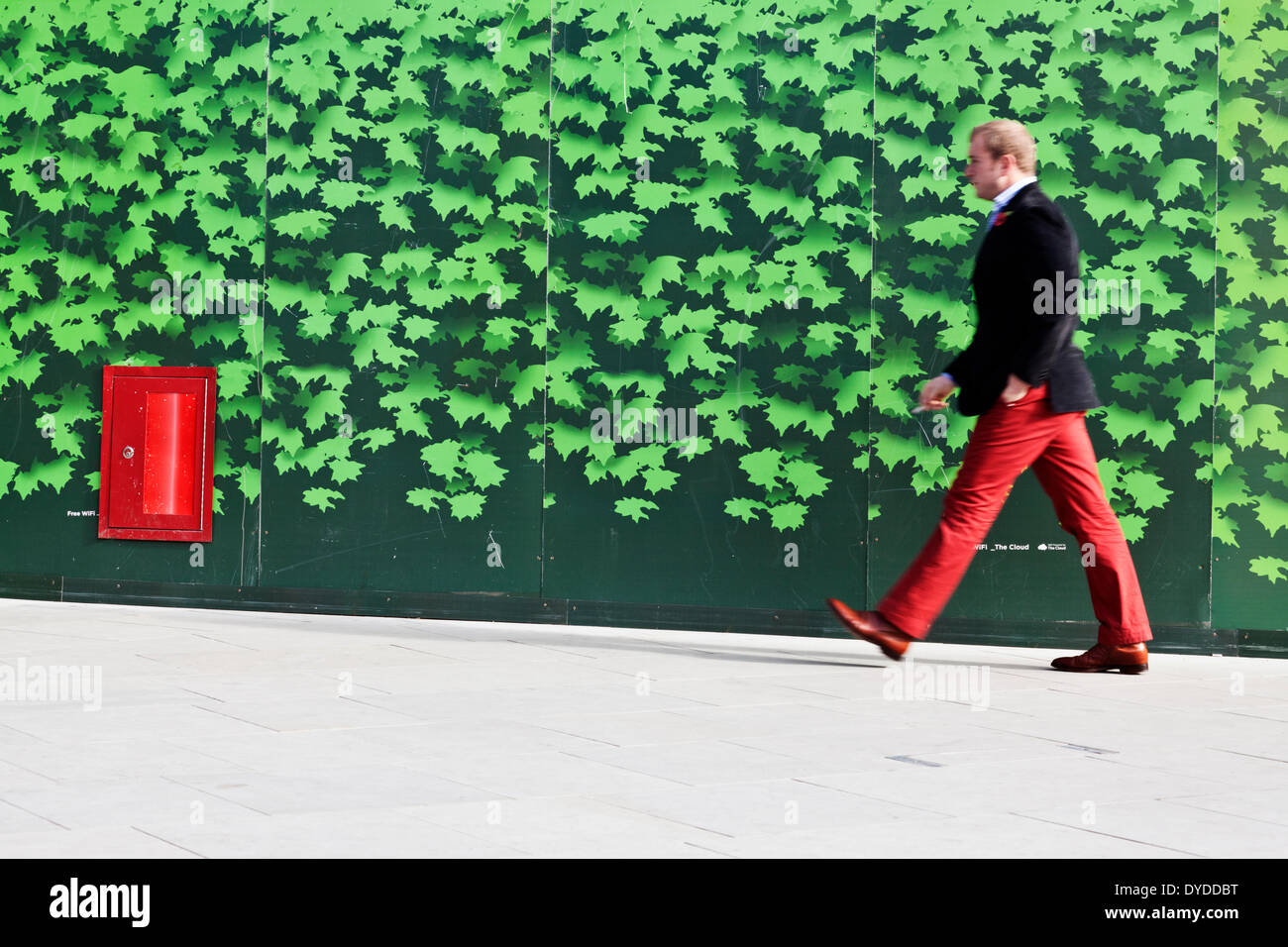 A man in red trousers and shoes walks by architectural hoardings. Stock Photo