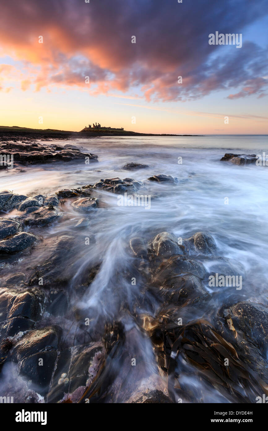 Sunset over Dunstanburgh Castle with waves breaking over the rocks in the foreground. Stock Photo