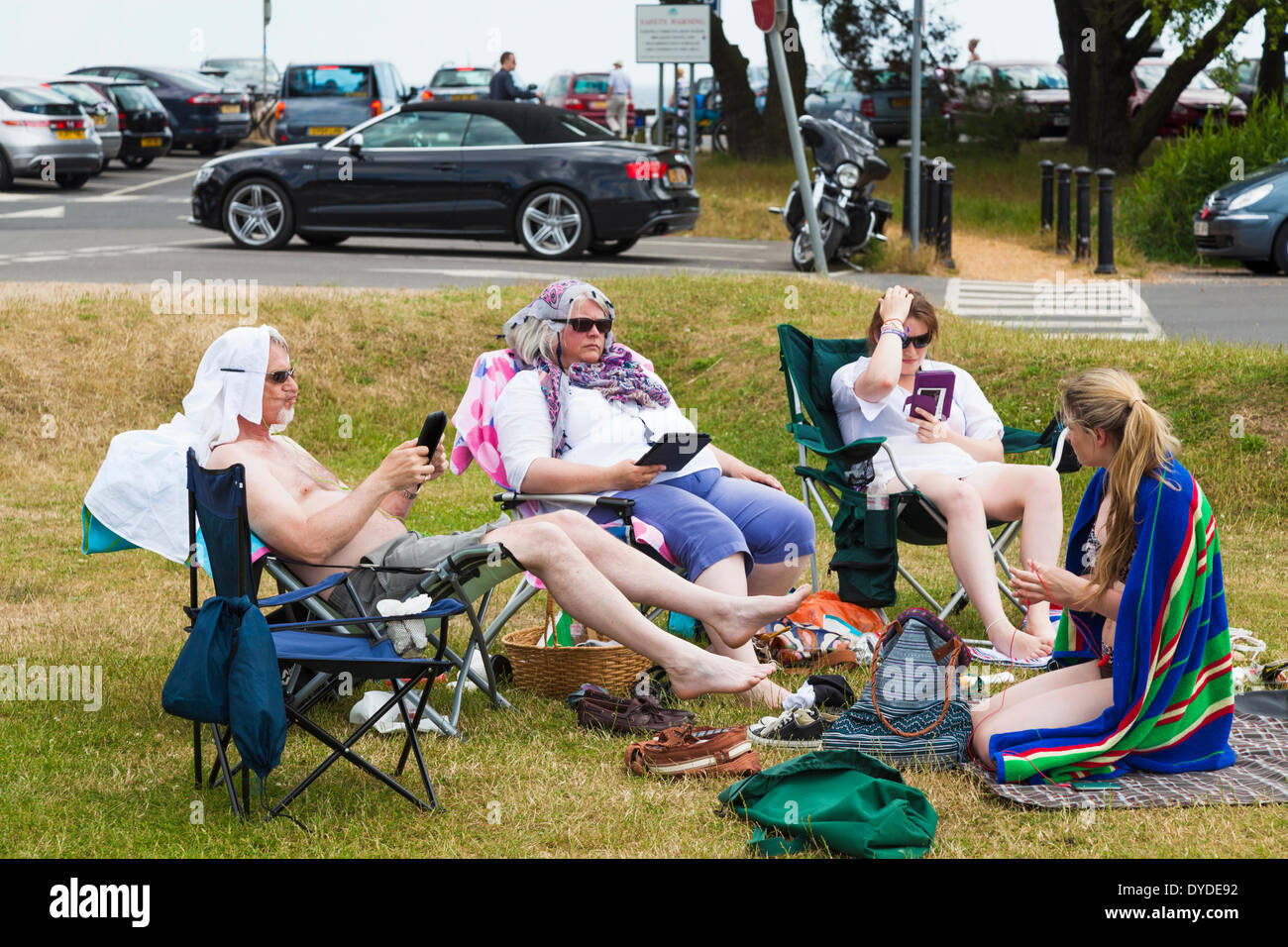 Family group sitting on grass by a car park and using e readers. Stock Photo