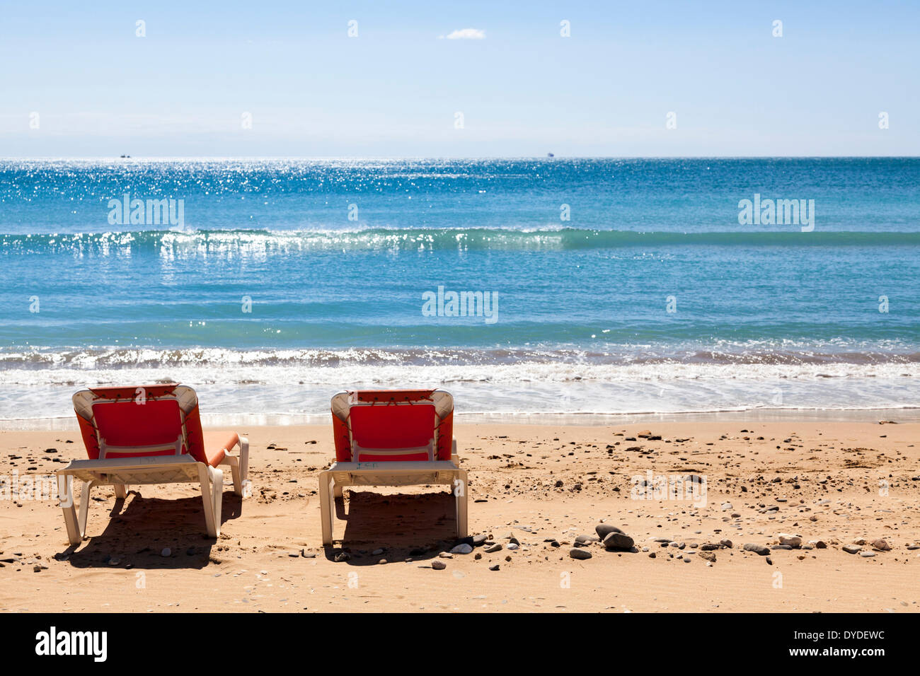 Two unoccupied red sun loungers on beach by sea. Stock Photo